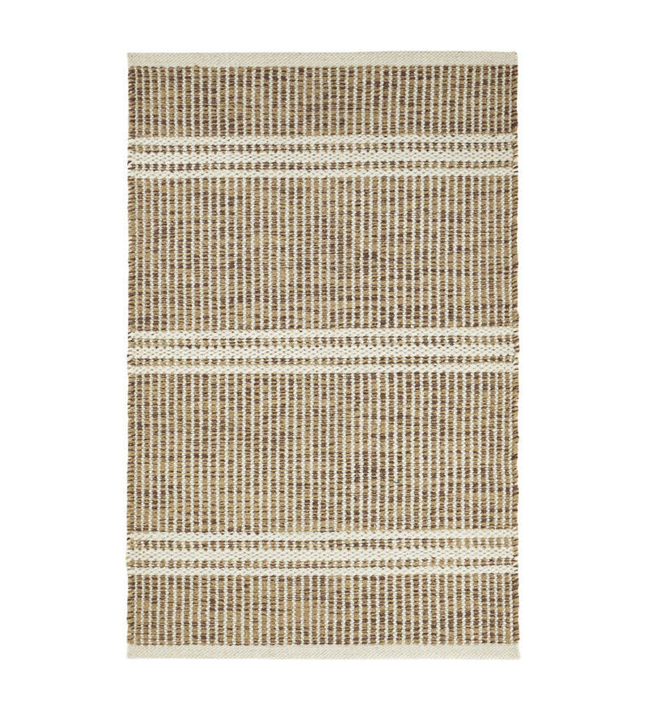 Malta Natural Woven Wool Rug displayed with a lounge chair, throw pillow, and plant. 