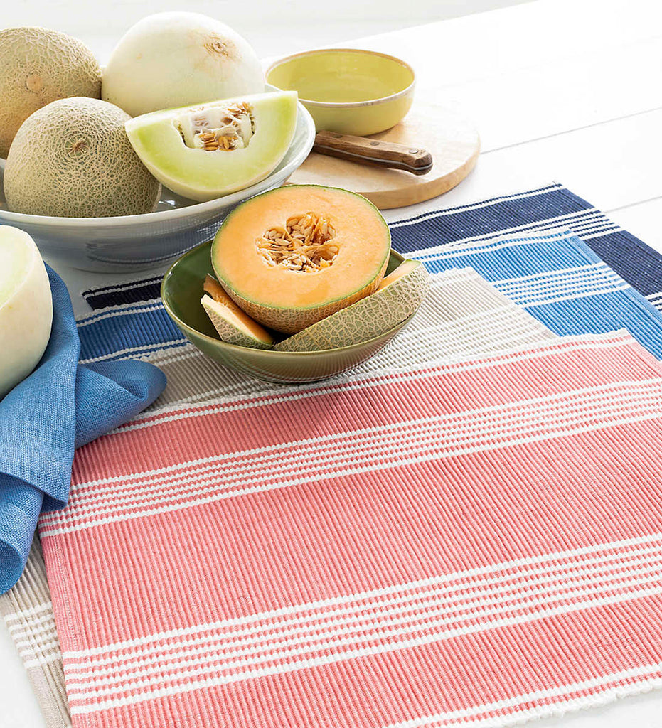 Tabletop display including: Bistro Stripe Placemats in Coral, Platinum, French Blue, and Indigo, alongside bowls of fresh cantaloupe.